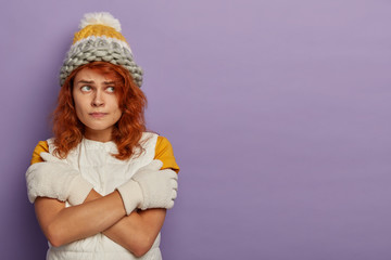 Beautiful redhead woman in wool knitted hat, white gloves and vest, crossed arms over body, trembles from cold, looks with unhappiness, has walk during frosty weather, isolated on purple wall