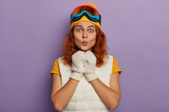 Surprised ginger female snowboarder keeps lips rounded, wears gloves, snowboarding mask, enjoys sliding on mountain during ski resort, looks directly at camera, isolated over purple background
