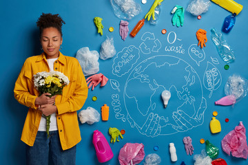 Tender curly haired female holds beautiful bouquet, has closed eyes, being eco friendly, stands against symbolic drawn Earth and plastic garbage, involved in nature cleaning. Consumption, recycling