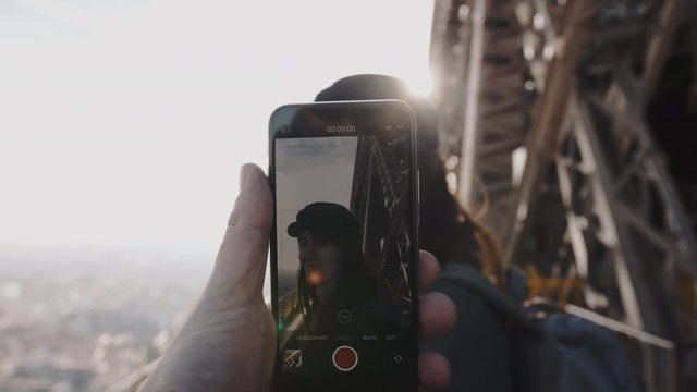 Male hand is holding smartphone filming happy smiling woman on top of Eiffel Tower. Romance and technology slow motion.