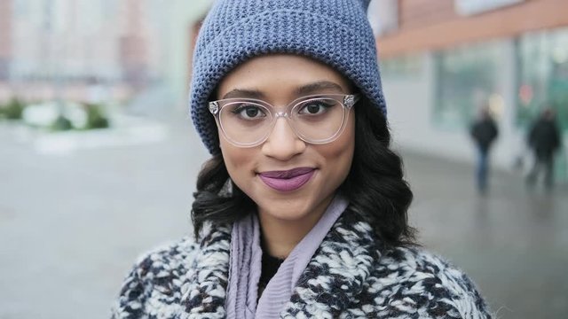 Close-up portrait of beautiful mixed race woman. Young woman in glasses looking at camera and smiling. Female in hat and coat outdoors in 4K, UHD