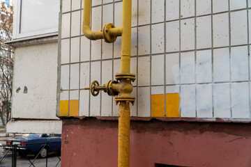Wall of building and branched vertical yellow gas pipe with valve. Valve with flywheel on the conduit allowed to distribute fuel gas and turn it off if needful. Wall with white tiles. Horizontal photo