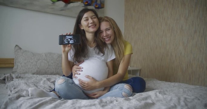 Pregnant multinational lesbian couple shows at an ultrasound image of his baby on the phone. LGBT