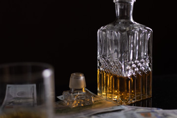 A crystal decanter with whiskey, next to an inverted crystal cork from it, hundred-dollar bills and...