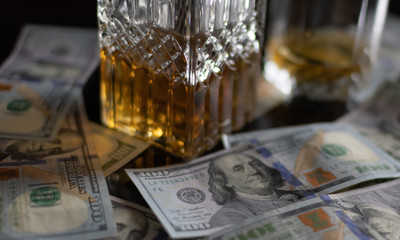 Hundred dollar bills are scattered on a black glass table. In the background is a blurry glass with whiskey and a crystal decanter with whiskey. On a black glass table