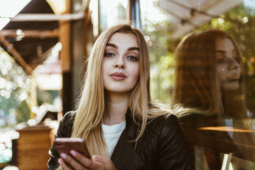 Cute blonde looks down at the camera, holds a phone, sits in a street cafe