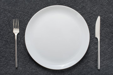 White empty plate with fork and knife on a grey tablecloth, top view.