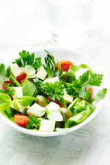 Tasty and healthy salad with feta cheese, green olives, cherry tomatoes and fresh herbs on white wooden background. Close up.