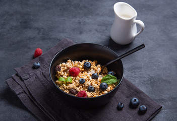 sweet muesli with berries in black plate with milk on dark table.