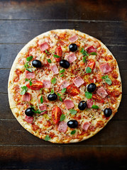 Pizza with cherry tomatoes, ham, black olives and fresh herbs. Home made food. Concept for a tasty and hearty meal. Brown wooden background. Top view. 