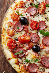 Pizza with sausage, cherry tomatoes, black olives and fresh herbs. Home made food. Concept for a tasty and hearty meal. Brown wooden background. Top view. 