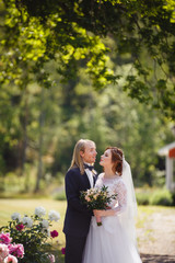 Young Bride and Groom couple in a summer garden on background of Swedish traditional house. Tender holding each other. Redhead woman and blonde man with long hairs. Young family near peony bush
