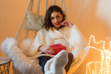 Beautiful young woman at home, opening red gift box, smiling. Warm light and festive cozy...
