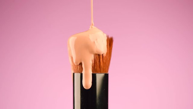 Makeup liquid foundation pouring on make-up contouring brush, closeup. Foundation beauty facial cosmetics, tool for perfect make up. Dripping bb cream or concealer, over pink background. 4K slow mo