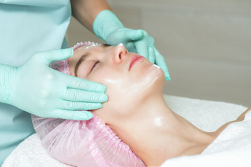 Cosmetologist's hands in gloves applying a white cream to the woman face in the spa beauty salon close up.