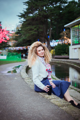 Portrait of smiling beautiful curly blonde woman in white knitted pullover and blue skirt sitting near pond with reusable coffee cup in lightened decorated park. Autumn, winter walking time. Vertical.