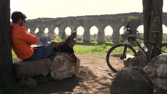 Cyclist with bike sitting on a rock under a tree on a videocall with his laptop in front of ancient roman aqueduct. Young attractive athletic man with orange sportswear and backpack in parco degli