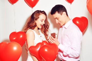 Man makes present to his lovely sweetheart girl. Lover's valentine day. Valentine Couple. Boy gives to his girlfriend jewelry. Wedding ring. Propose background red balloons hearts. Happy smile girl.