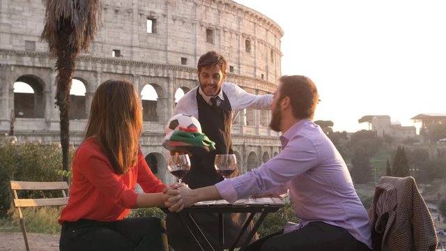 Elegant waiter serving football with italian flag to man of romantic young couple sitting at restaurant table in front of colosseum in rome at sunset the girl is annoyed