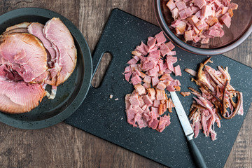 Black plate stacked with holiday ham, black cutting board with ham slices and scraps, kitchen knife, glass bowl