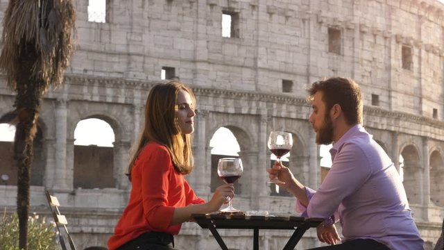 Romantic couple drinking and toasting with cup of red wine sitting at restaurant table in front of colosseum in rome at sunset