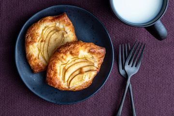Fresh individual apple tarts on a black plate with forks, mug of milk, purple cloth background