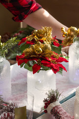 Woman making Christmas crafts with painted jars and poinsettias in her home. Wearing a red plaid shirt and a red plaid bow. 