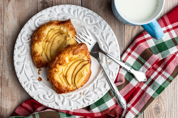 Fresh individual apple tarts on a white plate with forks, cup of milk, red, green, and white cloth napkin, wood background