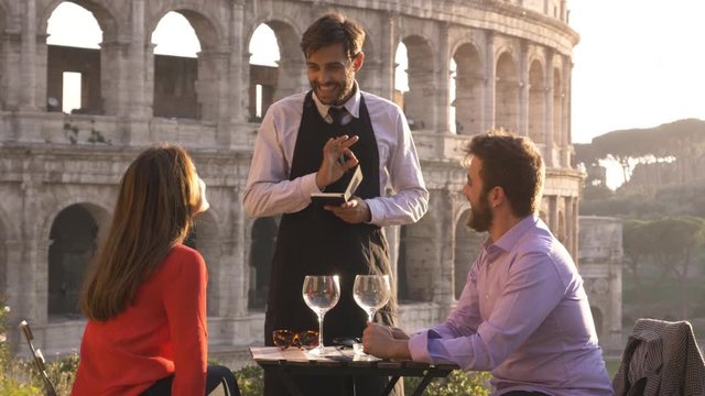Romantic elegant couple sitting at restaurant table in front of colosseum in rome at sunset ordering food and drinks with the waiter