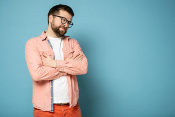 Cute bearded man with glasses, stands with folded arms and smiles good-naturedly, with copy space.