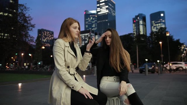 Two young pretty girls are sitting on suitcases in city park in evening, waiting. Female with ginger hair is sad, looking at apple watch on her wrist. Women are talking, discussing something.