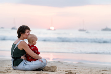 Naklejka premium Mom and baby enjoy the sunset at sea together in an embrace. Harmony and peace, mother kisses the child.