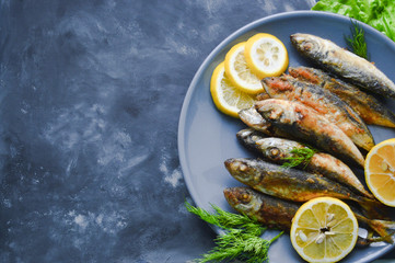 Fried horse mackerel istavrit fish skewered on a plate with lemon on the gray-black concrete background. Top view on the table with arugula's leaves.Healthy food concept, mediterranean diet.