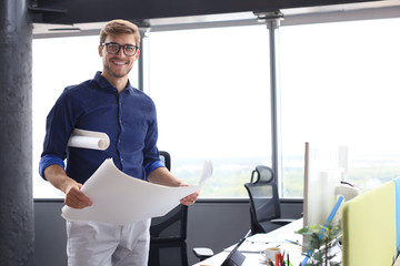 Confident young business man in shirt examining blueprint while standing against a window at office.