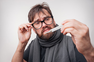 Flu season and colds. Sick young man looks at a thermometer. Neutral light background.