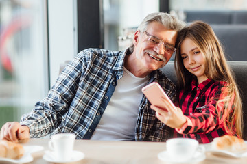Fototapeta premium Cute stylish granddaughter wearing red shirt make a selfie by phone with grandfather and croissant, having breakfast together.