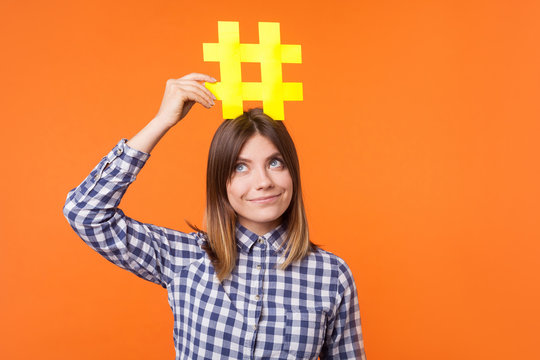 Popularity in social media. Portrait of funny brunette woman wearing checkered casual shirt holding large big yellow hashtag sign on head as crown. indoor studio shot isolated on orange background