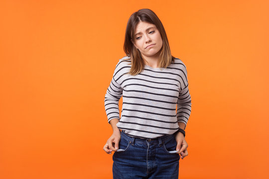 Portrait Of Upset Poor Young Woman With Brown Hair In Long Sleeve Striped Shirt Standing, Showing Empty Pockets In Denim, Having No Money, Bankrupt. Indoor Studio Shot Isolated On Orange Background