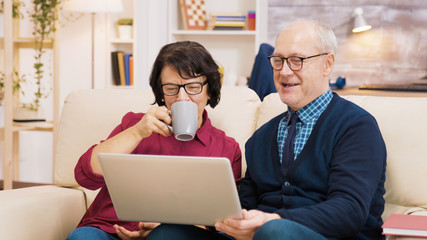 Elderly age couple sitting on sofa holding laptop during a video call.