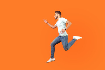 Fototapeta na wymiar Side view of joyous brunette man with beard in sneakers and denim outfit running in air, hurrying for discounts, empty copy space for advertising. indoor studio shot isolated on orange background