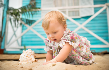 Charming smiling little girl in a floral dress sits on the sand against the background of a blue country house. Concept of walking outdoors for little children