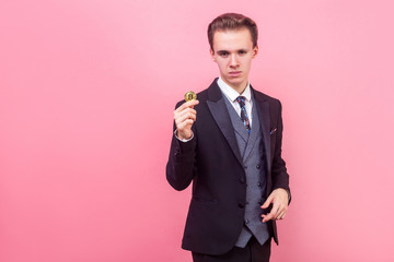 Portrait of smart confident businessman in elegant suit and with stylish haircut holding golden bitcoin, looking serious, copy space for cryptocurrency ad. indoor studio shot isolated, pink background