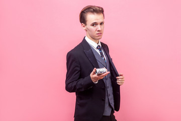 Portrait of arrogant rich businessman in elegant suit and with stylish haircut showing bunch of dollars on camera, offering money or boasting earnings. indoor studio shot isolated, pink background