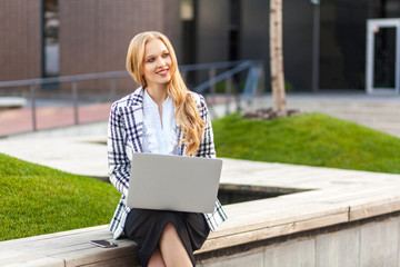 Portrait of elegant intelligent and extremely beautiful businesswoman with blond hair in plaid jacket sitting on bench working on laptop, looking inspired and satisfied with business idea, outdoors