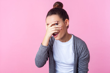 Portrait of adorable curious brunette teen girl with bun hairstyle in casual clothes peeking through fingers, looking embarrassed, pleasantly surprised. indoor studio shot isolated on pink background