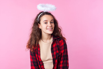 Portrait of sweet charming teenage girl with curly brunette hair wearing red plaid shirt and toy halo above head, smiling with kind angelic expression. indoor studio shot isolated on pink background