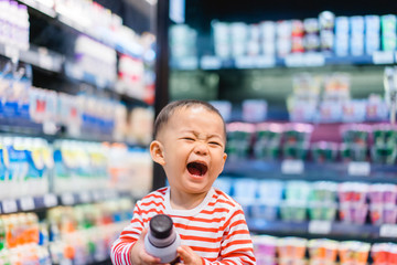 Funny little child holding chocolate milk and big laughing, little asian toddler boy, sitting in the trolley cart during family shopping in supermarket.