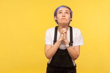 Please, I'm begging! Portrait of hipster woman with violet short hair in denim overalls looking up pleadingly and holding hands in prayer, begging forgiveness. indoor, yellow background, studio shot