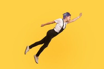 Superman. Portrait of enthusiastic ambitious hipster girl with violet short hair in denim overalls flying like superhero in air, feeling free, striving up for success. yellow background, studio shot
