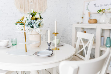 Stylish kitchen in white, pastel colors. Style minimalism. Vase with flowers, white table, candles,glasses, plates,dishes. Trendy interior with white furniture, table, brick walls. loft apartment